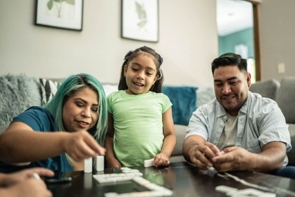 Mother and father sitting on the floor in the living room playing dominoes with a young daughter.